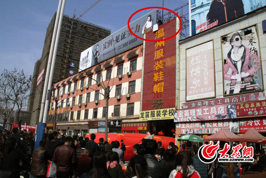 讨薪男子坐在楼顶广告架上，手持白色塑料板（图中红圈处）